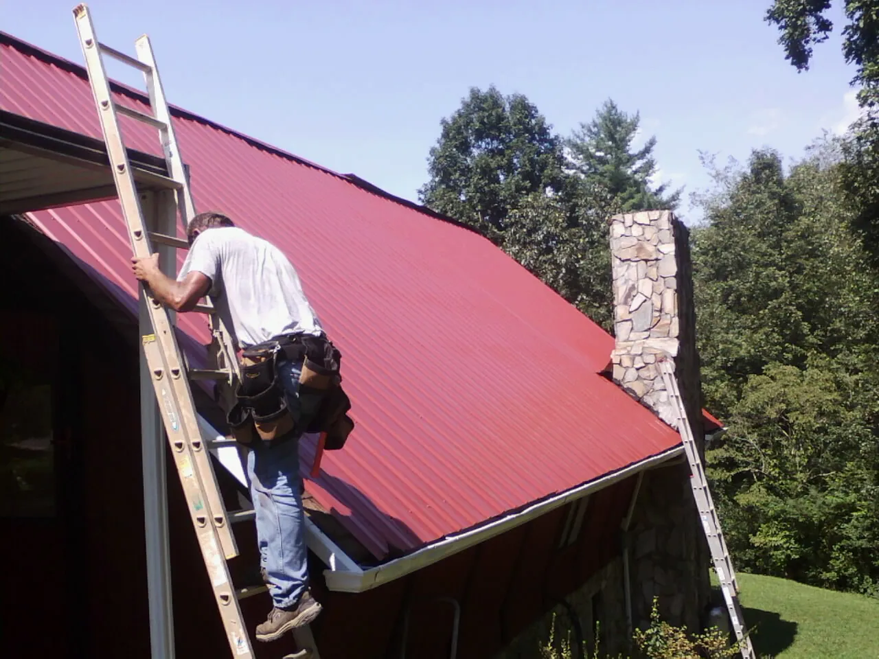 Roofer on a ladder beside a red metal roof and stone chimney — Soffit & Fascia Repair by Roofing Brothers in Belleville